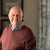 Portrait of happy retired senior man standing at home near window. Satisfied old man in casual clothes looking at camera and smiling while standing near the window. Positive and confident elderly enjoy his retirement at care facility.
